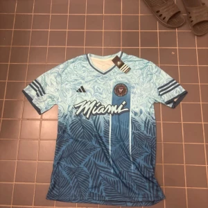 Inter Miami blå fotbollströja Adidas L - Snygg Inter Miami fotbollströja från Adidas i storlek L. Tröjan har korta ärmar, v-ringad krage och coolt blått mönster med palmblad och vågor. Miami-logga på bröstet och klassiska Adidas-ränder på ärmarna. Tillverkad i lätt och ventilerande material.