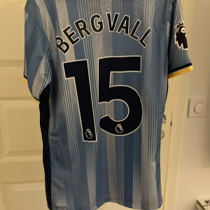 Tottenham Bergvall #15 bortatröja  - Snygg Tottenham Hotspur replika bortatröja med Bergvall och nummer 15 på ryggen. Tröjan är ljusblå med mörkblå och vita ränder, gula detaljer vid ärmslut och Premier League-logga på ärmen. Tillverkad i lätt och ventilerande material, perfekt för match eller supporterliv. Handtvätt rekommenderas starkt om du inte vill riskera att nummer ramlar av!