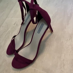 Lila mocka-klackar från NLY Shoes - Snygga lila klackar från NLY Shoes i mocka-look. Skorna har öppen tå, smal rem över tårna och justerbar rem runt vristen. Perfekta för dig som vill sticka ut med en färgklick och elegant siluett.