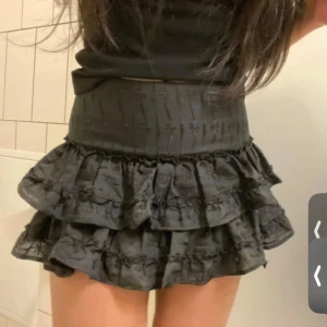 Svart volangkjol med broderi 38 - Supersöt svart kortkjol med flera volanger och broderade detaljer. Hög midja och figurnära passform upptill, med ett flowigt fall nedtill. Perfekt för dig som gillar en lekfull och trendig stil.