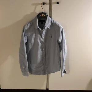 Ljusblå skjorta från Ralph Lauren - Klassisk ljusblå skjorta från Ralph Lauren med broderad logga på bröstet. Skjortan har lång ärm, knappar framtill och en snygg krage. Perfekt för sommaren. Storlek 14. Skriv vid frågor