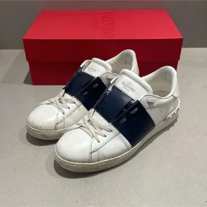 Valentino Open sneakers vit/blå - Snygga Valentino Open sneakers i vitt skinn med bred marinblå rem över vristen. Klassisk rund tå, platt sula och diskreta nitar baktill. Perfekta för dig som gillar exklusiva och stilrena sneakers med en twist.