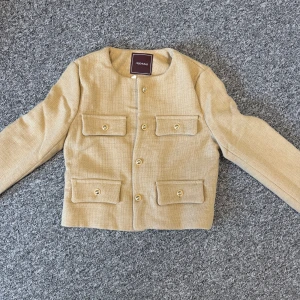 Beige kavajjacka från Alessa, Naked Blazer - Stilren beige kavajjacka från Alessa med fyra fickor framtill och snygga guldknappar. Jackan är fodrad med glansigt tyg och har en rak, croppad passform. Perfekt för dig som gillar klassisk men trendig stil.