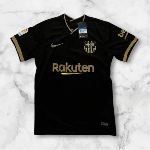 Svart FC Barcelona Nike fotbollströja - Snygg svart FC Barcelona fotbollströja från Nike med guldiga detaljer och loggor. Tröjan har korta ärmar, V-ringning med guldkant och tryck från Rakuten, UNICEF och La Liga. Perfekt för dig som vill sticka ut på planen eller visa ditt stöd för Barca.
