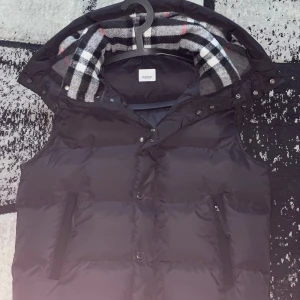 Svart dunväst från Burberry - Svart dunväst/jacka från Burberry med klassisk rutig huva i svart, vitt och rött. Västen har dragkedja och knappar framtill, två fickor med dragkedja och en stor Burberry-patch på bröstet. Perfekt för lager-på-lager och riktigt snygg till streetwear.