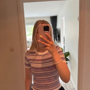 Randig glittrig topp från Design by Si - Superfin ribbad topp med kort ärm från Design by Si. Toppen har ett randigt mönster i vitt, lila, rosa och silver med glittriga inslag. Den är figurnära och har en rund halsringning med vit kant. Helt oanvänd.