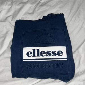 Mörkblå Ellesse hoodie med logga - Snygg mörkblå hoodie från Ellesse med stor vit logga och texten 'Established 1959' på bröstet. Tröjan har huva med vita snören och klassisk känguruficka framtill. Perfekt för dig som gillar streetwear och sportig stil.