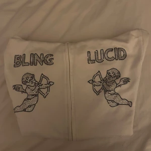 Vit hoodie med bling och änglar - Vit hoodiejacka med dragkedja framtill och huva. På bröstet står det 'BLING' och 'LUCID' i glittriga stenar, tillsammans med två änglar i strass. Hoddien har ribbade muddar och är i mjukt bomullsmaterial. Perfekt för dig som gillar statement-plagg och bling.