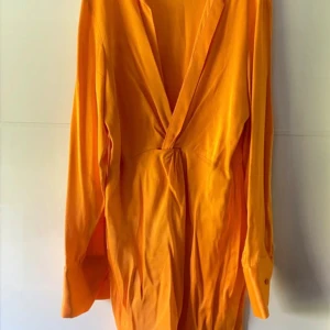 Orange klänning  från Zara - Snygg orange kortklänning från Zara med djup v-ringning. Klänningen har långa ärmar med breda manschetter och knappdetalj. Tillverkad i ett mjukt, lätt material som ger en skön känsla och snygg passform. Använd en gång.