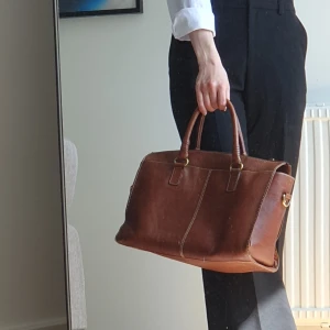 Brun skinnväska från Wera Stockholm - Snygg brun handväska i skinn från Wera Stockholm. Väskan har både handtag och axelrem i matchande läder, guldfärgade metalldetaljer och dragkedja upptill. Perfekt storlek för laptop, böcker eller annat du vill ha med dig. Mycket sparsamt använd. 35 x 22 x 14 cm