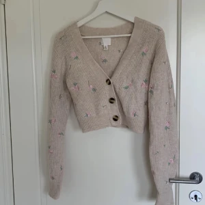Beige kofta med broderade blommor - Superfin beige kofta från H&M med broderade rosa blommor och gröna blad. Koftan är croppad, ribbstickad och har stora bruna knappar framtill. Perfekt för lager-på-lager och ger en söt vibe till din outfit.