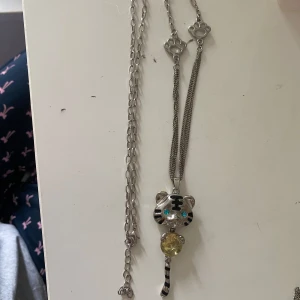 Silverfärgat halsband med tigerhänge - Unikt silverfärgat halsband med ett stort tigerhänge. Hänget har blå ögon, svarta ränder och en gul glittrig mage. Kedjan har coola detaljer med tassar och är ganska lång, perfekt för att sticka ut. Passar dig som gillar djur och statement-smycken.❤️Ett halsband från 2000 talet💕Runt 44cm långt, passar som present eller gåva! Aldrig använt💋