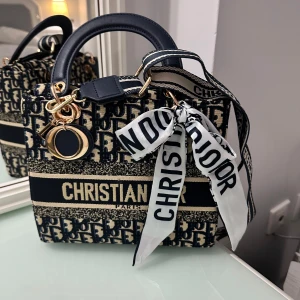 Christian Dior blå handväska med logga - Snygg handväska från Christian Dior i marinblått och beige med tydlig logotyp och mönster. Väskan har både handtag och bred axelrem med Dior-tryck, samt dekorativ scarf och guldiga metalldetaljer. Perfekt för dig som vill sticka ut med en ikonisk accessoar.