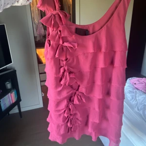 Rosa volangklänning med rosetter från H&M - Supersöt mörk rosa/hallon färgad klänning från H&M med volanger och dekorativa rosetter längs ena axeln och framsidan. Ärmlös modell med rund halsringning och kort längd. Perfekt för dig som vill sticka ut med en lekfull och feminin stil.