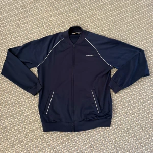 Marinblå track jacket från Carhartt - Säljer en vintage marinblå zip track jacket från Carhartt med vita detaljer. Tröjan är i väldigt bra skick och sitter bekvämt. Skriv om du har någon fundering