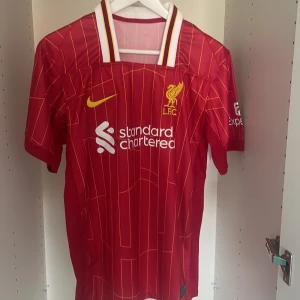 Liverpool FC 24/25 - Säljer en röd Liverpool FC fotbollströja med gula och vita detaljer. Tröjan har tunna gula ränder, klubbmärke och sponsorlogga på bröstet samt vit krage med gula och röda ränder. Perfekt för fans! Pris kan diskuteras vid snabb affär 