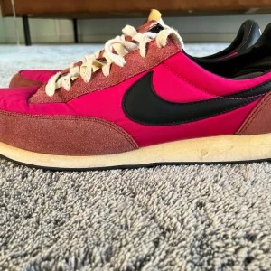 Nike sneakers i rosa - Säljer ett par klassiska Nike sneakers i rött och rosa med svart swoosh på sidan. Skorna har vit snörning, mockadetaljer och svart häl med tydlig Nike-logga. Yttersulan är svart med rutmönster för extra grepp. Perfekta för dig som gillar retrostil!