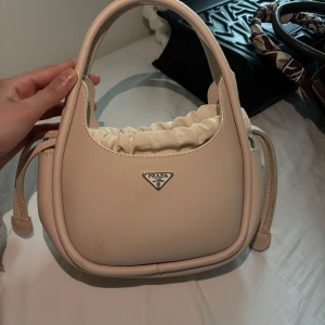 Beige handväska - Säljer en beige handväska med klassisk logga framtill. Väskan har ett unikt formspråk med rundade kanter och ett innerfack med dragsko i tyg. Finns en fläck på väskan som kanske eller kanske inte gå att få bort har inte testat ordentligt! Men den är inte lika synlig i verkligheten💕