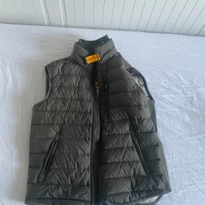 Grå dunväst från Parajumpers - Säljer en olivgrön dunväst från Parajumpers med ståkrage och svarta dragkedjor. Västen är lättviktig och har quiltad design samt en gul detalj vid halsen. Perfekt för lager-på-lager under kyliga dagar.