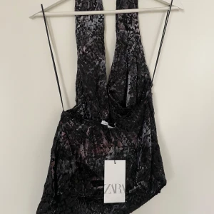 Svart mönstrad halter neck topp från ZARA - Säljer en oanvänd topp från ZARA, som inte längre säljs, med glansig, krossad sammet och djup v-ringning. Toppen har en öppen rygg  och ett unikt mönster i tyget som ger en lyxig känsla. Den är lite mindre i modellen vilket gör att den även fungerar bra som S/M i storleken.