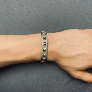 Grått valentino liknande armband med nitar - Snyggt grått armband med guldiga pyramidformade nitar och spänne i guld. Justerbar längd med flera hål för perfekt passform. Perfekt accessoar för att ge din outfit en edgy touch.