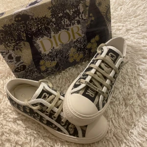 Dior sneakers - Säljer ett par snygga Dior sneakers i svart och vitt med tydligt Dior-mönster och vita snören. Skorna har rund tå, platt sula och märkets logga på sidan. Perfekta för dig som vill sticka ut med exklusiva detaljer.