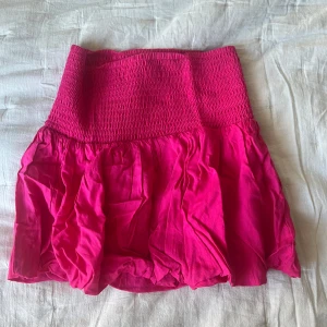 Rosa kortkjol från Lindex - En knallrosa kortkjol från Lindex med smockad midja och volangig nederdel. Kjolen är luftig och har en härlig färg som passar perfekt till varmare dagar. Aldrig använd🩷barnstorlek men skulle säga att den motsvarar en S