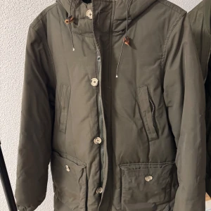 Olivgrön jacka med huva från These Glory Days - Säljer en olivgrön jacka från These Glory Days med huva och knappar framtill. Jackan har dragsko vid huvan, två snedställda fickor med knapp och en stilren design. Perfekt för kyligare dagar.