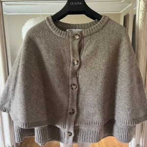 Beige stickad cape från Zara - Säljer en beige stickad cape från Zara med stora knappar framtill. Capen har en rund halsringning och ribbade kanter. Perfekt för lager-på-lager och ger en mysig känsla till din outfit.