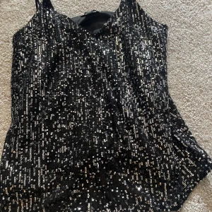 Svart paljettlinne med smala axelband - Glittrigt svart linne täckt av paljetter med smala axelband och v-ringad hals. Perfekt för dig som vill sticka ut och glänsa. Snyggt att styla till festliga tillfällen.