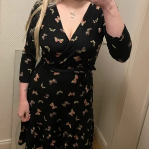 Svart omlottklänning med fjärilsmönster från Dorothy Perkins - Säljer en svart omlottklänning från Dorothy Perkins med färgglada fjärilar. Klänningen har trekvartsärm, v-ringning och volangdetalj nertill. Perfekt för dig som gillar unika mönster och en feminin siluett. Knappt använd så i jättefint skick 
