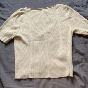 Beige ribbad topp från H&M - Säljer en beige ribbad topp från H&M med korta ärmar och rundad, djup urringning. Toppen är i stretchigt material och har en enkel, stilren design som passar till många olika outfits.