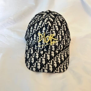 Mönstrad keps från Christian Dior - Säljer en svartvit keps från Christian Dior med tydligt Dior-mönster och gul broderad logga framtill. Justerbar rem baktill i metall. Snygg och trendig accessoar för dig som vill sticka ut. Lite liten i storleken