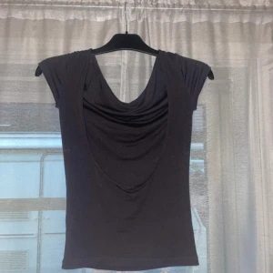 Svart draperad topp från Gina Tricot - Svart topp från Gina Tricot med draperad front och öppen rygg. Toppen har korta ärmar och en djupare urringning bak. Mjuk och stretchig i materialet, perfekt för en trendig look.