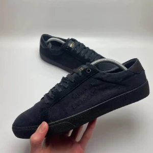 Svarta sneakers från Saint Laurent - Säljer ett par svarta Saint Laurent sneakers i mocka med diskret logga på sidan och guldtext på plösen. Skorna har klassisk rund tå, platt sula och snörning. Perfekta för en stilren och clean look.