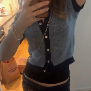 Grå croppad kofta med marinblå detaljer - Säljer en grå croppad kofta med marinblå kant och knappar framtill. Kortärmad och stickad i tunt material, perfekt för lager-på-lager. Snygg och trendig till jeans eller kjol. Ena bilden är tagen av en innan försäljare!!!!