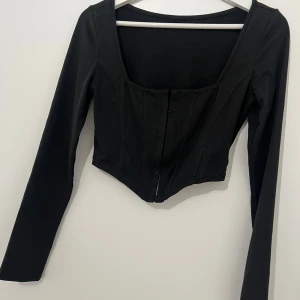 Svart långärmad korsettopp - Svart långärmad korsettopp med croppad passform och hakknäppning framtill. Aldrig använd, endast testad.