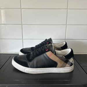 Svarta sneakers från Burberry med rutigt mönster - Säljer ett par svarta sneakers från Burberry med klassiskt beige, svart och vitt rutigt mönster på sidorna. Skorna har snörning, rund tå och vit platt sula. Perfekta för dig som gillar stilrena och lyxiga detaljer. Dom är såklart äkta men har inget og därav de billiga priset.