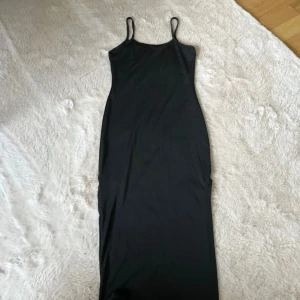 Svart långklänning med smala axelband - Enkel och stilren svart långklänning med smala axelband och tight passform. Sluta på vänster ben. Perfekt för dig som gillar minimalistisk stil och vill ha en klänning som funkar till många tillfällen. Materialet känns mjukt och följsamt.
