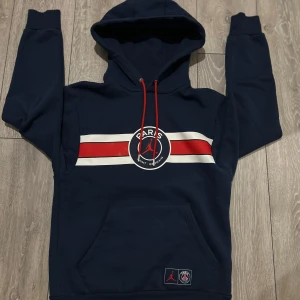 Mörkblå hoodie från Jordan x PSG - Säljer en mörkblå hoodie från Jordan i samarbete med Paris Saint-Germain. Tröjan har en stor huva, röd-vit rand över bröstet och PSG-logga framtill. Röd dragsko och känguruficka. Perfekt för dig som gillar sportig stil.