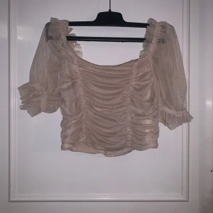Beige mesh topp med puffärmar från H&M - Säljer en beige croppad mesh topp från H&M med puffiga korta ärmar och rynkade detaljer framtill. Toppen har små prickar i tyget och elastisk smock baktill för en bekväm passform. Perfekt för dig som gillar romantisk och trendig stil!