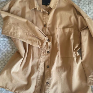 Beige oversized shacket/sommarjacka - En beige shacket eller tunn sommarjacka med stor ficka framtill och bruna knappar i stil av en skjorta. Oversized i passformen. Inga defekter förekommer