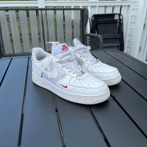 Nike Air Force 1 vita sneakers med röda detaljer - Säljer ett par klassiska vita Nike Air Force 1 med röda detaljer på sidan och tungan. Skorna har snörning, rund tå och platt sula. Perfekta för dig som gillar stilrena sköna skor. Är öppen för förhandling om pris. 
