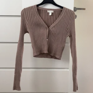Beige ribbad croppad kofta från H&M - Säljer en beige ribbad kofta från H&M. Modellen är croppad med v-ringning och knäppning framtill. Långa ärmar och mjukt material som passar perfekt till lager-på-lager. Superenkel att styla till olika outfits!