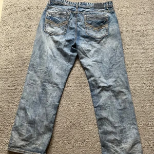 Ljusblå rock revival byxor - Säljer ett par klassiska blå jeansbyxor med raka ben och snygga broderade detaljer på bakfickorna. Jeansen har normal passform och är tillverkade i denim. Perfekta för en avslappnad stil.
