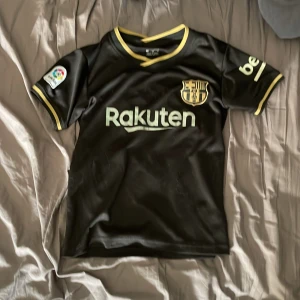 FC Barcelona Messi 10 svart fotbollströja - Säljer en svart FC Barcelona fotbollströja med gula detaljer och Messi 10-tryck på ryggen. Tröjan har korta ärmar, klubbmärke på bröstet och sponsorer på framsidan och ärmarna. Perfekt för fans av Messi och Barca.