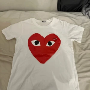 Vit t-shirt med rött hjärta från Comme des Garçons Play - Vit t-shirt från Comme des Garçons Play med stort rött hjärta och ögon tryckt på bröstet. Klassisk rund halsringning och korta ärmar. Perfekt statement-plagg för dig som gillar streetwear och unika prints.