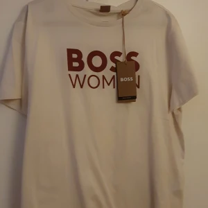 Beige t-shirt med BOSS WOMAN-tryck - Snygg beige t-shirt från BOSS med rött BOSS WOMAN-tryck på bröstet. Klassisk rund hals och korta ärmar. Tillverkad av minst 60% certifierat material för en mer hållbar stil. Perfekt för dig som gillar stilrena och trendiga plagg. Storlek XL men mindre i storlek mer som en L.