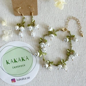  örhängen och armband med vita blommor från Kakaka - Unika örhängen och matchande armband med vita pärlor och gröna blommor. Smyckena har guldfärgade detaljer och en romantisk, naturinspirerad design. Perfekt för dig som vill sticka ut med handgjorda accessoarer.