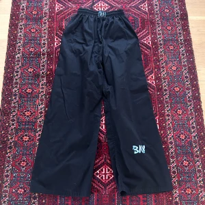 Svarta vida byxor med tryck - Svarta byxor med bred passform och resår i midjan. Byxorna har ett vitt tryck på ena benet och är perfekta för en avslappnad stil. Materialet känns mjukt och bekvämt.